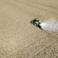 Zielony traktor z przyczepą rozsypuje nawóz na polu. To ważny etap prac polowych, który zapewnia prawidłowy wzrost roślin. Nikosar etykieta na przyczepie informuje o zawartości.