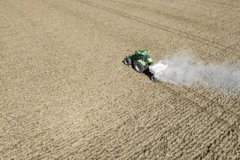 Zielony traktor z przyczepą rozsypuje nawóz na polu. To ważny etap prac polowych, który zapewnia prawidłowy wzrost roślin. Nikosar etykieta na przyczepie informuje o zawartości.